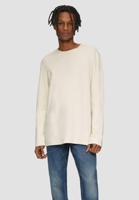 Langärmliges, cremefarbenes Baumwollhemd mit Rundhalsausschnitt und relaxed Fit, kombiniert mit blauen Jeans, vor einem neutralen Hintergrund.