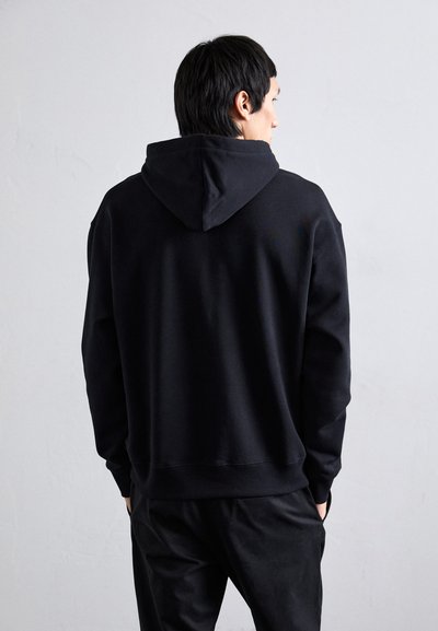 Schwarzer Kapuzenpullover mit Kängurutasche, Kordelzug und gerippten Bündchen. Der Stoff scheint weich mit einer glatten Textur zu sein und wird mit dunklen Hosen getragen.
