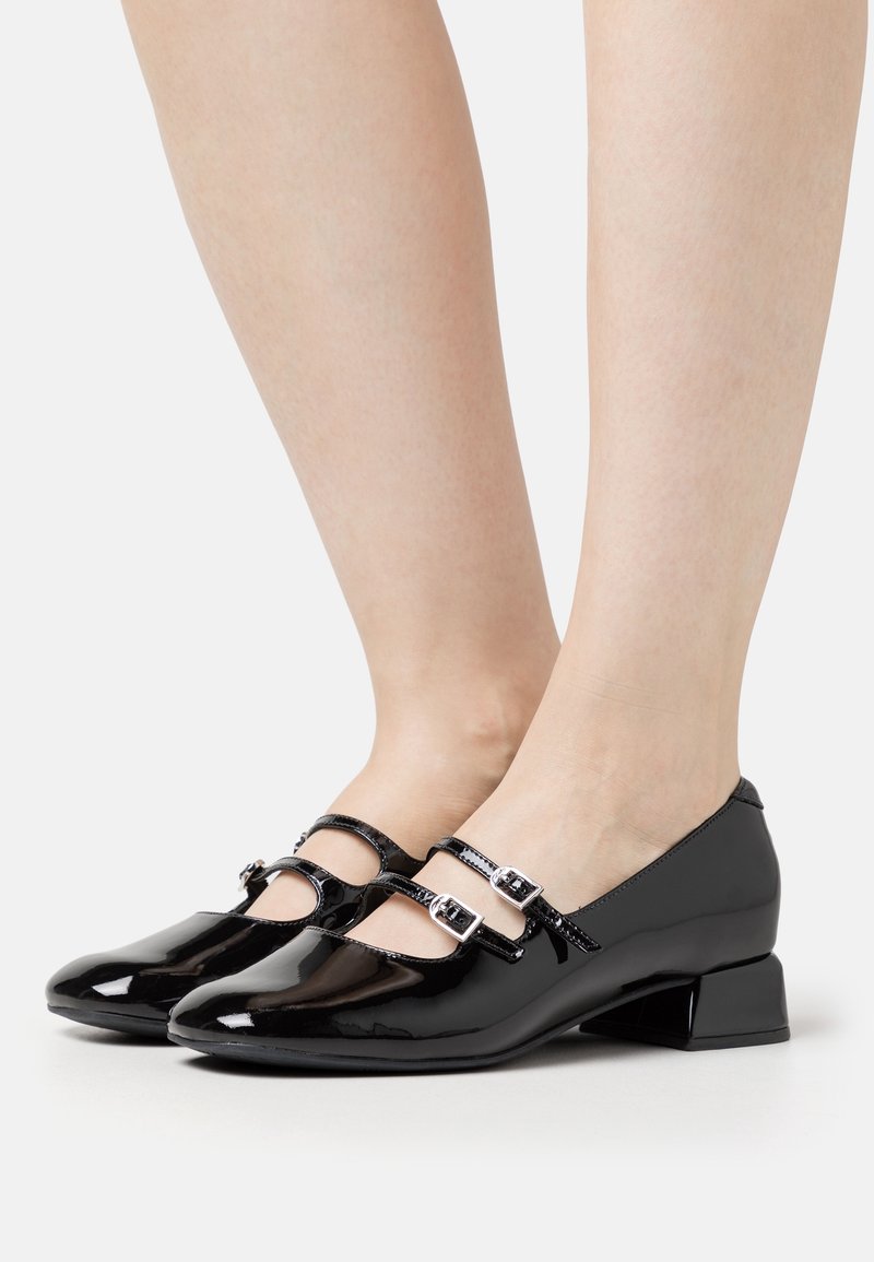 Jambes portant des chaussures Mary Jane en cuir verni noir brillant avec des brides à double boucle et des talons blocs bas sur fond blanc.