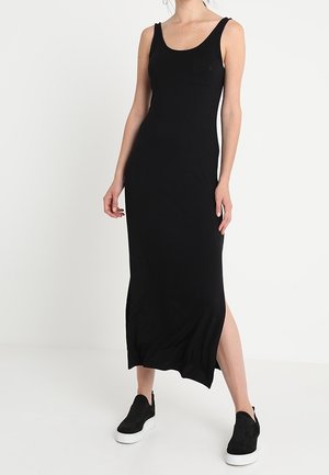 Mujer con vestido midi negro sin mangas con abertura lateral y zapatillas negras sin cordones con suelas blancas sobre un fondo blanco.