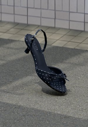 Schwarzer hochhackiger Sandale mit Knöchelriemen und weißen Punkten, verziert mit einer Schleife an der Spitze, aufrecht stehend auf einem grauen Gehweg in der Nähe einer gefliesten Wand.