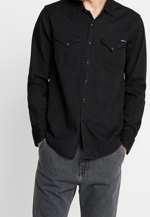 Mężczyzna w czarnej dżinsowej koszuli na guziki z kieszeniami na piersiach oraz szarych dżinsach, ręce w kieszeniach, na jednolitym jasnym tle.