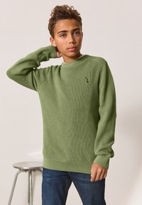 Vihreä ribbineuloksinen neulepaita pyöreällä kaula-aukolla, pitkät hihat ja pieni tunnus rinnassa. Päällä siniset farkut.