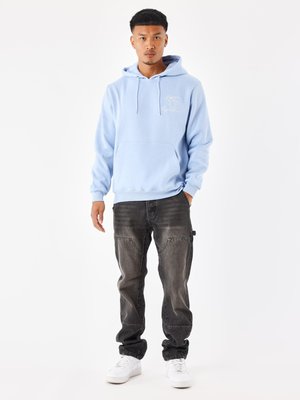 Haut à capuche bleu clair avec une poche avant et des détails brodés, associé à un jean noir déchiré et des baskets blanches, se tenant sur un fond uni.