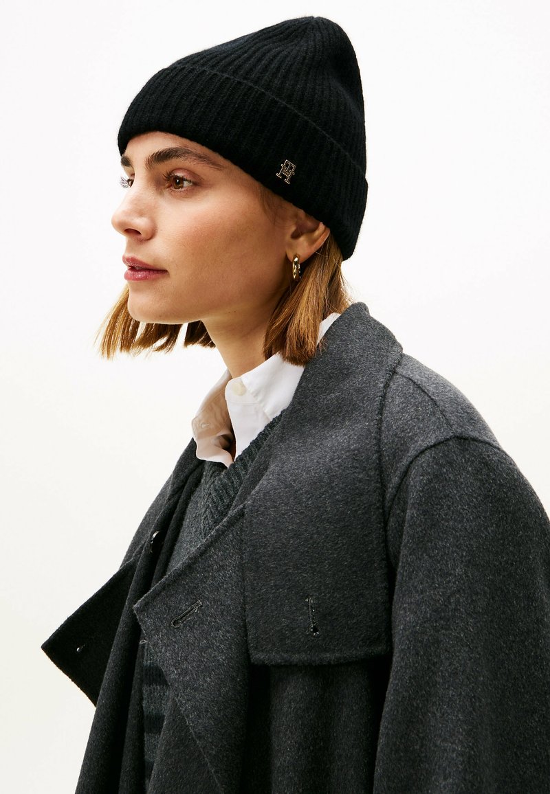 Black ribbed knit beanie paired with a dark gray wool coat, featuring a collar, layered knit sweater, and a white button-up shirt underneath.