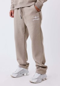 Beige sweatpants with elastic waistband and drawstring; features side pockets and white logo; paired with gray sneakers. Smooth texture.