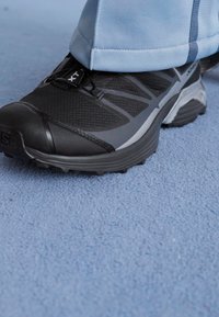 Black athletic shoe with a textured upper, mesh panels, and grey accents. Features a rugged sole for traction and stability.