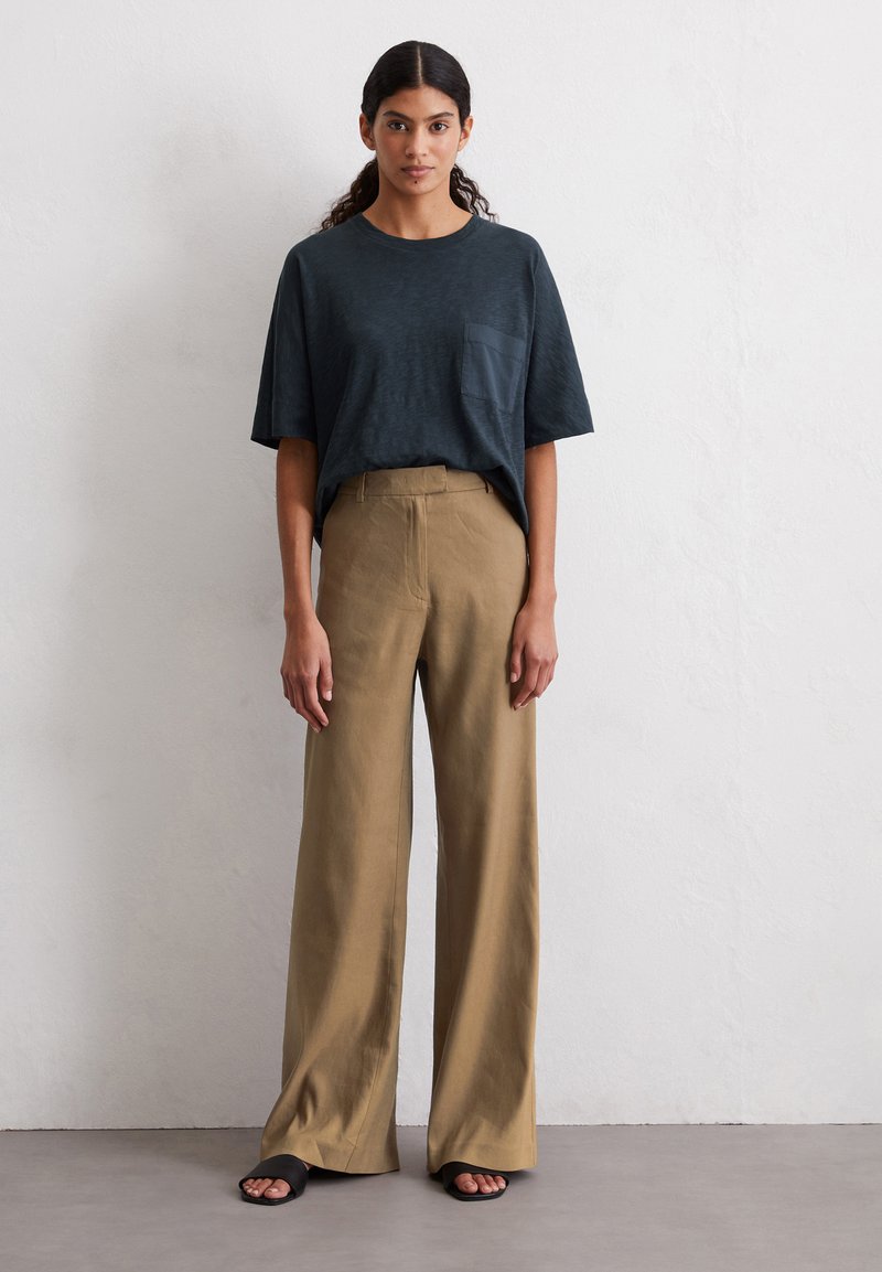 Dark blue short-sleeve t-shirt with pocket, paired with high-waisted, wide-leg beige trousers, and black slide sandals on a neutral background.