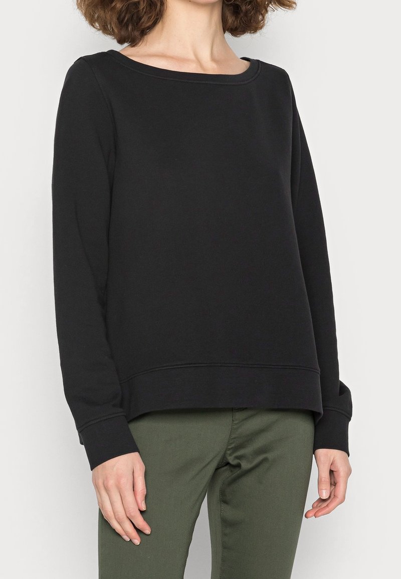 Svart långärmad sweatshirt med rund halsringning, tillverkad av mjukt material, med ribbade ärmslut och avslappnad passform, i kombination med gröna byxor.