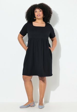 Vestido negro de manga corta con escote cuadrado, cintura recogida y bolsillos laterales. Combinado con zapatos planos grises claros. Textura de tela suave.
