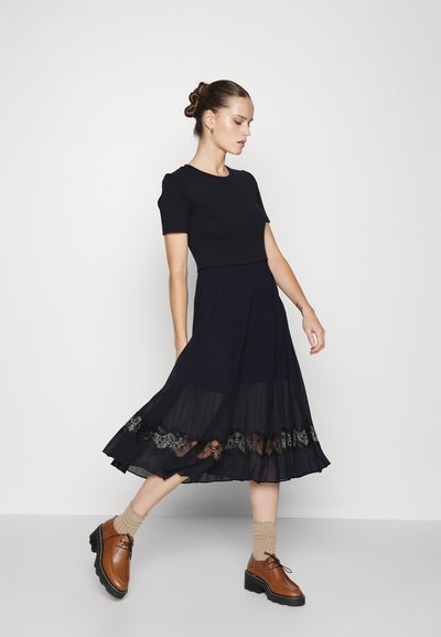 Vestido negro de manga corta con parte superior ajustada, falda fluida y adornos de encaje en el dobladillo. Combinado con zapatos marrones y calcetas beige.