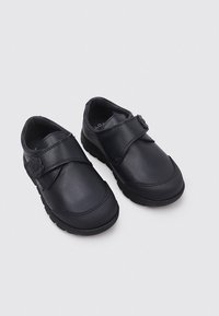 Zapatos de cuero negro con punta redondeada, cierre de tira de Velcro y suela de goma texturizada. Adornados con un detalle en forma de flor en la tira.