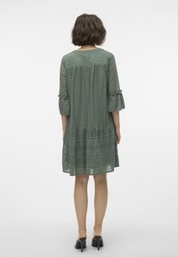 Robe en dentelle verte avec des manches trois-quarts, design superposé et accents froncés, présentant des motifs complexes et une coupe ample.