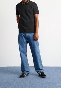 Svart texturerad kortärmad polotröja, blå vida denimjeans och svarta sneakers med vita ränder. Neutral bakgrund.