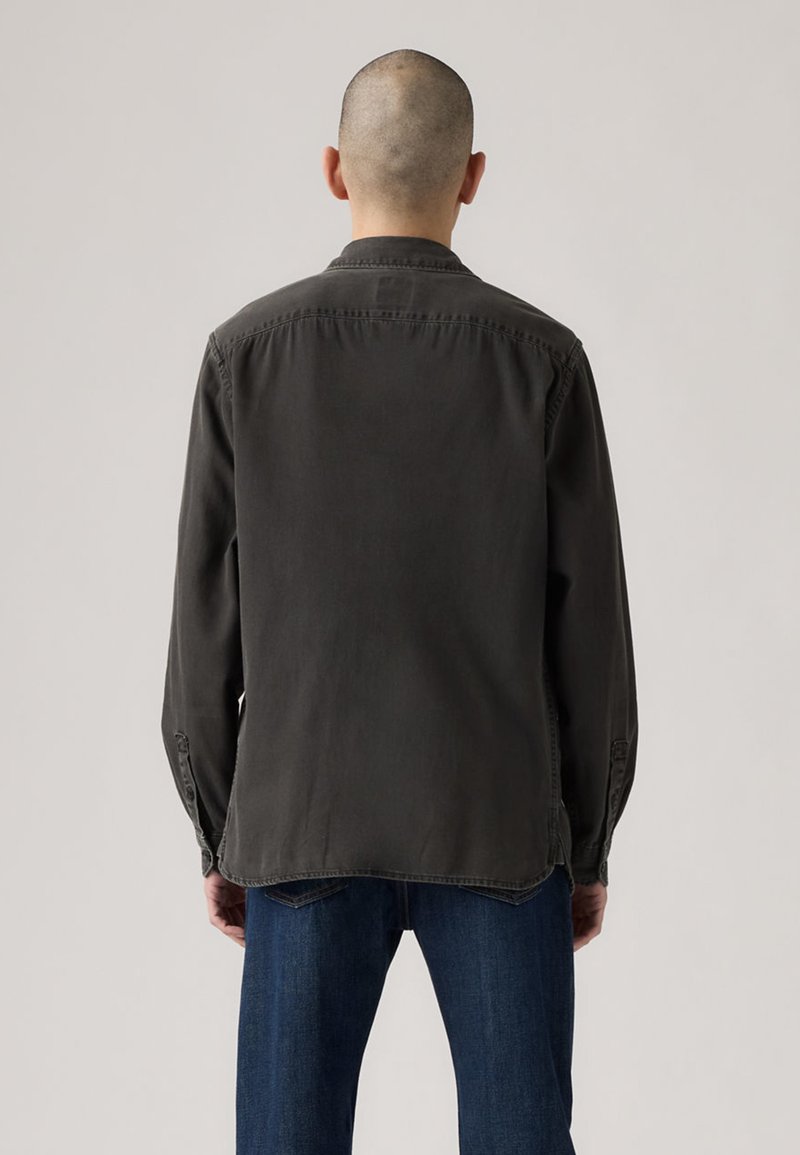 Chemise à manches longues gris foncé avec col, poignets à boutons et ourlet droit, portée par une personne vêtu de jeans bleus.