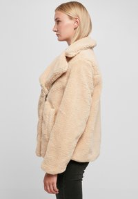 Beige nepsheerling jasje met een grote kraag, voorrits en zijzakken; heeft een losse pasvorm en een gestructureerd oppervlak.