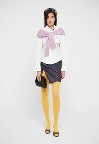 Wit overhemd met lange mouwen, paarse trui om de nek geknoopt, marineblauwe gestreepte rok, gele panty en zwarte handtas. Bruine peeptoe-hakken.