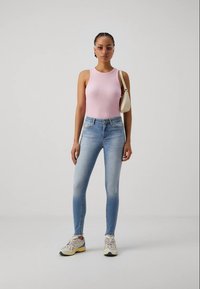 Top de alças canelado rosa claro, jeans skinny azuis e tênis cinzas com detalhes amarelos. Modelo segura uma pequena bolsa de creme.