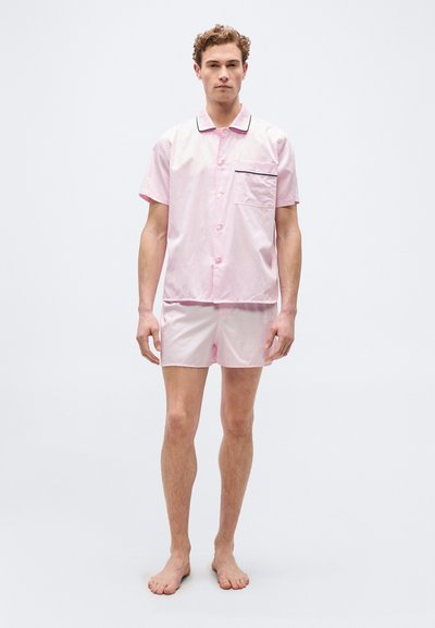 Άνδρας μοντέλο στέκεται ξυπόλητος, φορώντας ανοιχτό ροζ κοντομάνικο πουκάμισο με μαύρο περίγραμμα και ταιριαστό ροζ σορτς, σε απλό λευκό φόντο.