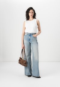Top sin mangas blanca y ligera combinado con unos vaqueros acampanados de cintura alta en azul claro. La modelo sostiene un pequeño bolso marrón de hombro. Texturas suaves, estilo casual.