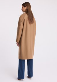 Kobieta z długimi brązowymi włosami, ubrana w długi beżowy płaszcz, niebieskie szerokie jeansy oraz szpilki w leopardzi wzór, zwrócona bokiem z głową obróconą w lewo.