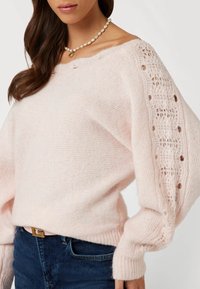 Rosa Strickpullover mit zarten Spitzenakzenten an den Ärmeln, oversized Schnitt und geripptem Saum. Weiche Textur und gemütliches Erscheinungsbild.