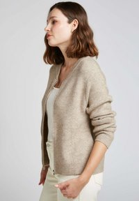 Cardigan beige en tricot doux, avec une fermeture éclair à l'avant, des poignets côtelés et une coupe décontractée avec une texture subtile.