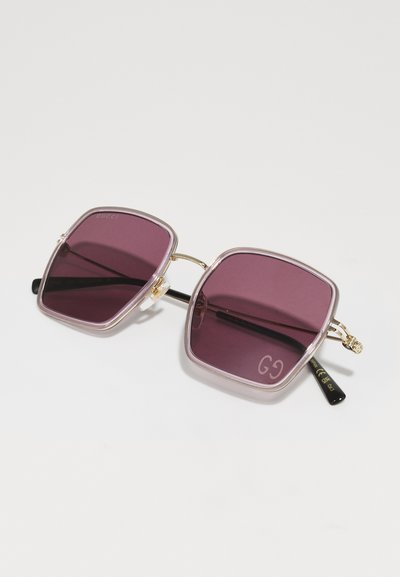 Gucci kvadratiniai saulės akiniai su purpuriniais objektyvais, skaidriais rėmeliais ir auksinėmis detalėmis. Yra juodas šventykla su prekės ženklo detalėmis.