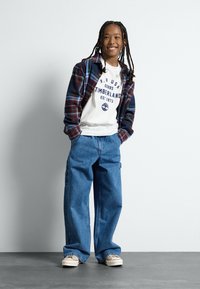 Conjunto infantil que incluye una camisa de franela a cuadros en colores oscuros, una camiseta gráfica blanca y unos vaqueros azules holgados con bolsillos.