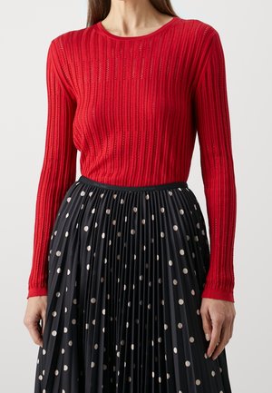 Kobieta ubrana w czerwony, długi rękaw sweter z pionowymi paskami oraz czarną plisowaną spódnicę w białe kropeczki.