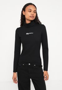 Crni dugi rukav turtleneck od glatkog materijala, s bijelim printanim logom na prsima. U kombinaciji s crnim trapericama.