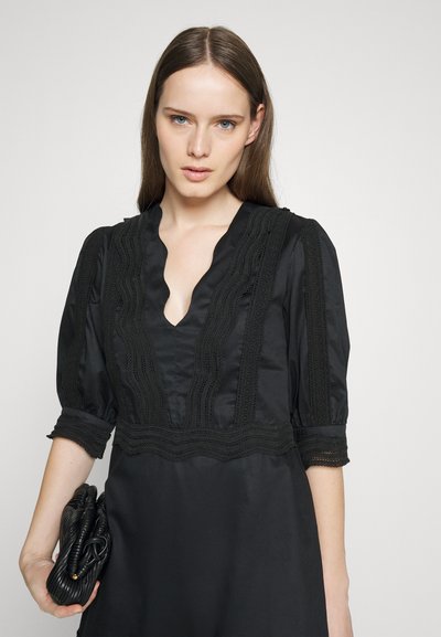 Γυναίκα με ίσια καστανά μαλλιά, ντυμένη με ένα μαύρο φόρεμα με δαντέλα, κρατώντας μια διπλωμένη μαύρη τσάντα με φόντο απλό.