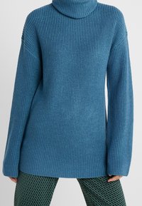 Pull en maille bleue côtelée avec un col cheminée haut, une coupe oversize, des manches longues et des fentes latérales, associé à des leggings verts à motifs.