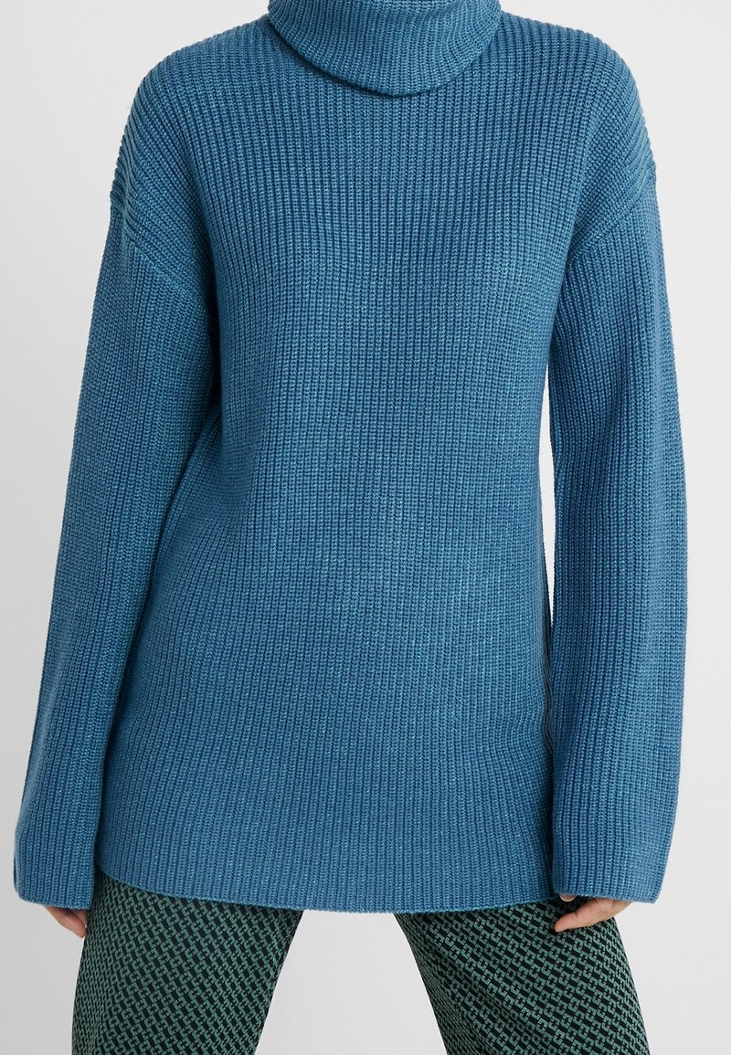 Pull en maille bleue côtelée avec un col cheminée haut, une coupe oversize, des manches longues et des fentes latérales, associé à des leggings verts à motifs.