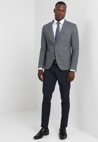 Grauer melierter Blazer mit Reverskragen, einreihiger Knopfverschluss und zwei Fronttaschen. Getragen mit einem weißen Hemd und marineblauen Hosen. Schwarze Schuhe.