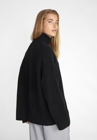 Pull en maille noire à col roulé avec une coupe ample, des poignets et un ourlet côtelés, et un tissu textur é. Modèle présenté en profil contre un fond blanc.