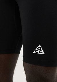 Κοντινή λήψη μαύρου αθλητικού σορτς με λευκό τριγωνικό λογότυπο ACG στο μηρό, που φοριέται από άτομο με σκούρο χρώμα δέρματος.