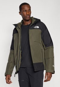 Veste isolante verte et noire avec fermeture éclair à l'avant, capuche et logo sur la poitrine. Elle présente une finition mate et un design épuré.