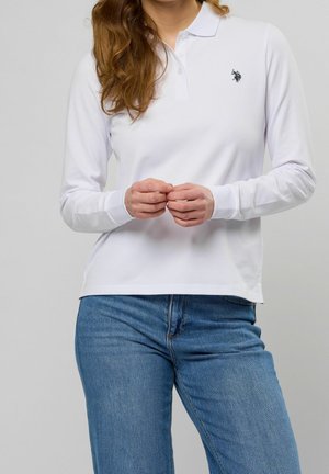 Kobieta w białej koszulce polo z długim rękawem z małym czarnym logo i niebieskich dżinsach, stojąca na tle jednolitego szarego tła.