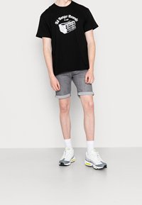 Camiseta gráfica negra con texto impreso en blanco, pantalones cortos de denim gris con puños enrollados y zapatillas grises con acentos en neón. Calcetines blancos visibles.