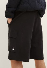 Osoba v černých kraťasích do kolen s boční kapsou a malým bílým logem, s rukou v kapse, doplněných tmavě prošívanou bundou.