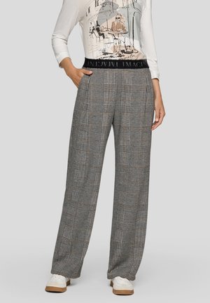 Frau trägt karierte, hoch taillierte, weit geschnittene Hose mit schwarzem elastischem Bund und weißen Turnschuhen, Hand in der Tasche, grauer Hintergrund.