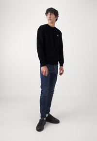 Sweatshirt noir à col rond, jeans en denim, baskets noires. Design simple avec un petit logo sur la poitrine. Texture lisse.