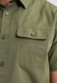 Camisa de algodón verde oliva con botón en el frontal, mangas cortas, bolsillo en el pecho y logo impreso en el bolsillo. Textura suave.