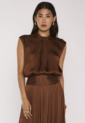 Giovane donna con lunghi capelli scuri che indossa un top marrone lucido senza maniche e una gonna plissettata abbinata, accessoriata con orecchini a cerchio dorati e anelli.
