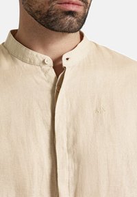 Camicia di lino beige con colletto alla cinese, caratterizzata da una chiusura frontale con bottoni e un sottile dettaglio ricamato "AIX" sul tessuto.