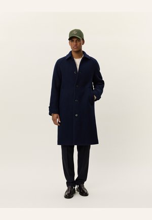 Navy blue wool overcoat with a classic design, featuring a button front, two side pockets, and a notched collar. Worn over a light shirt.