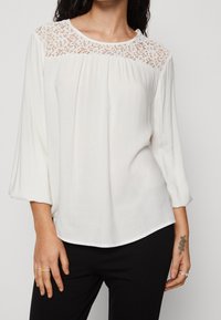 Vit blus med spetsdetaljer vid yolken, långa ärmar och en avslappnad passform. Slätt tyg med en lätt lyster. Matchad med mörka byxor.