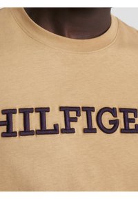 Närbild av en beige skjorta med stora djärva mörkblå broderade bokstäver som stavar "HILFIGER" över bröstet.
