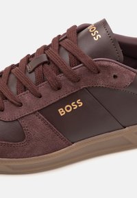 Brauner Sportschuh aus Wildleder und Leder, mit strukturierter Oberfläche, goldfarbenem Logo und runden Schnürsenkeln. Gummisohle mit profilierter Lauffläche.